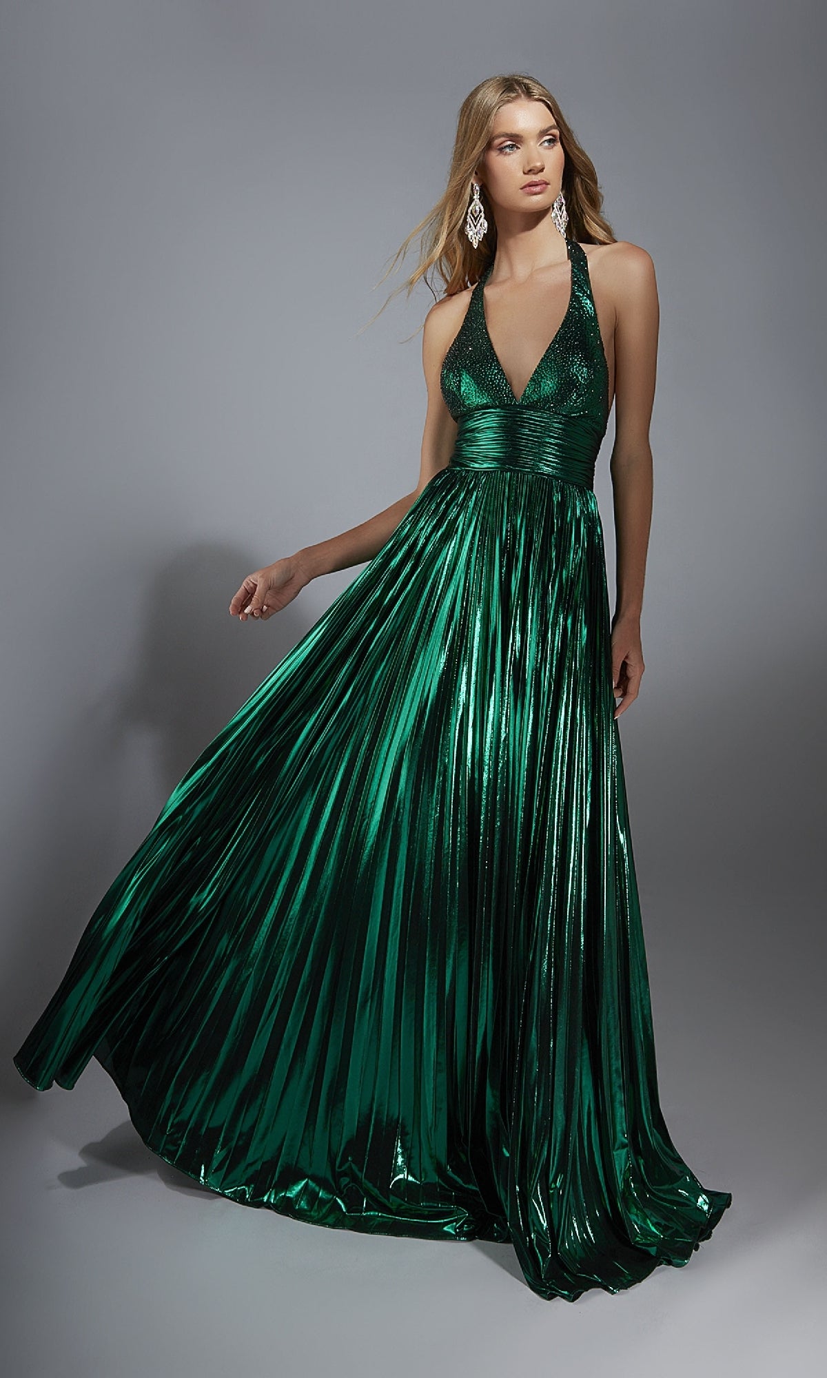 A woman models the Alyce 61792 long prom dress—shiny metallic emerald green with a V-neck halter neckline and pleated, flowing skirt—standing against a plain gray background.